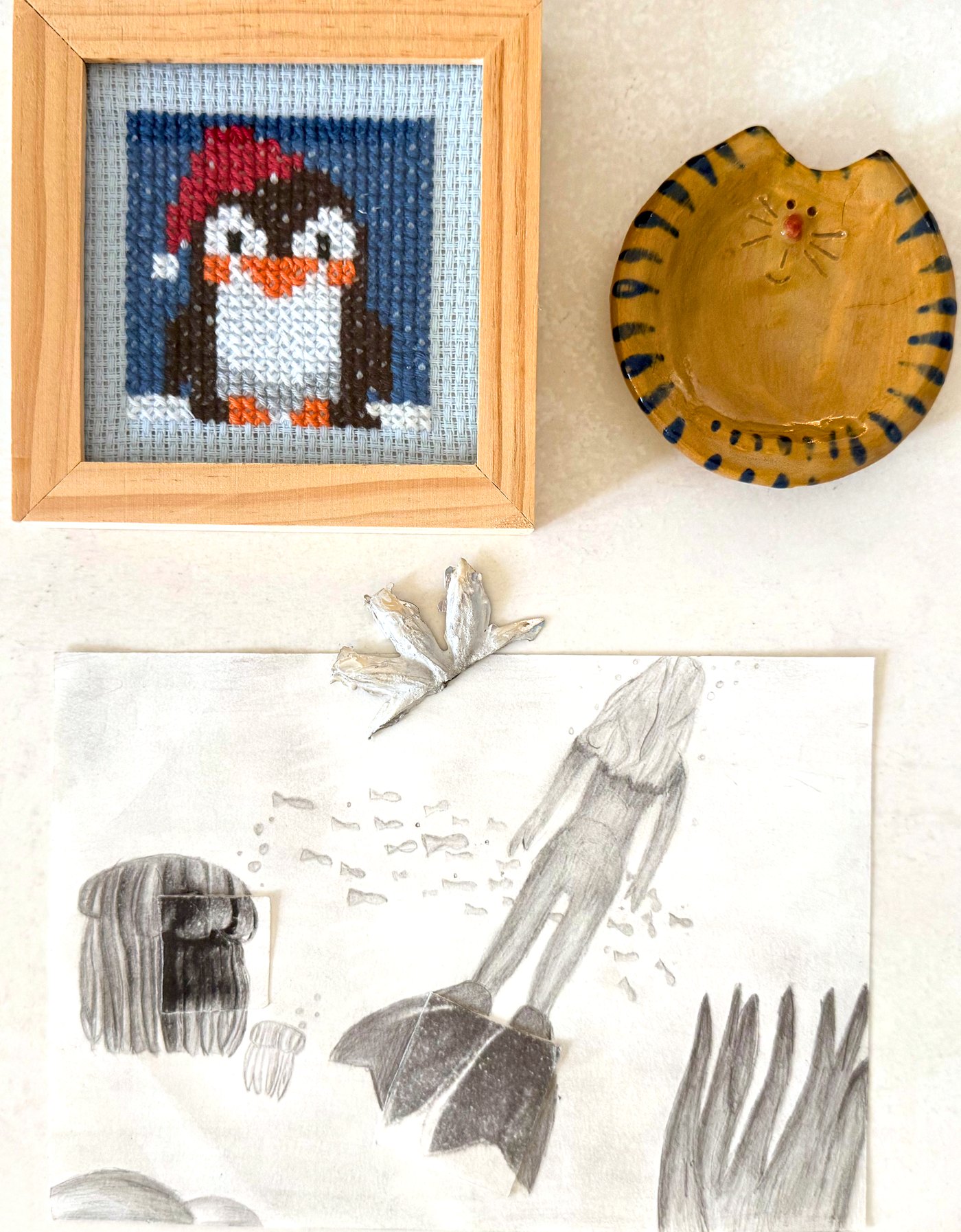 Four childhood creations photographed together on an ivory surface: a framed cross-stitch penguin, a ceramic cat dish, a pressed metal flower and a pencil drawing of a diver underwater.